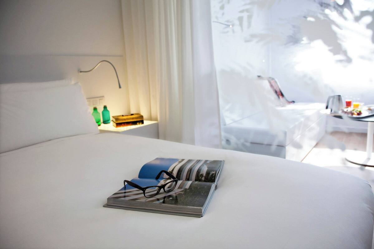 Ein ordentlich bezogenes Bett mit Buch und Brille darauf, ein Nachttisch mit Flaschen, und Blick auf einen Balkon mit Stuhl und Tisch.