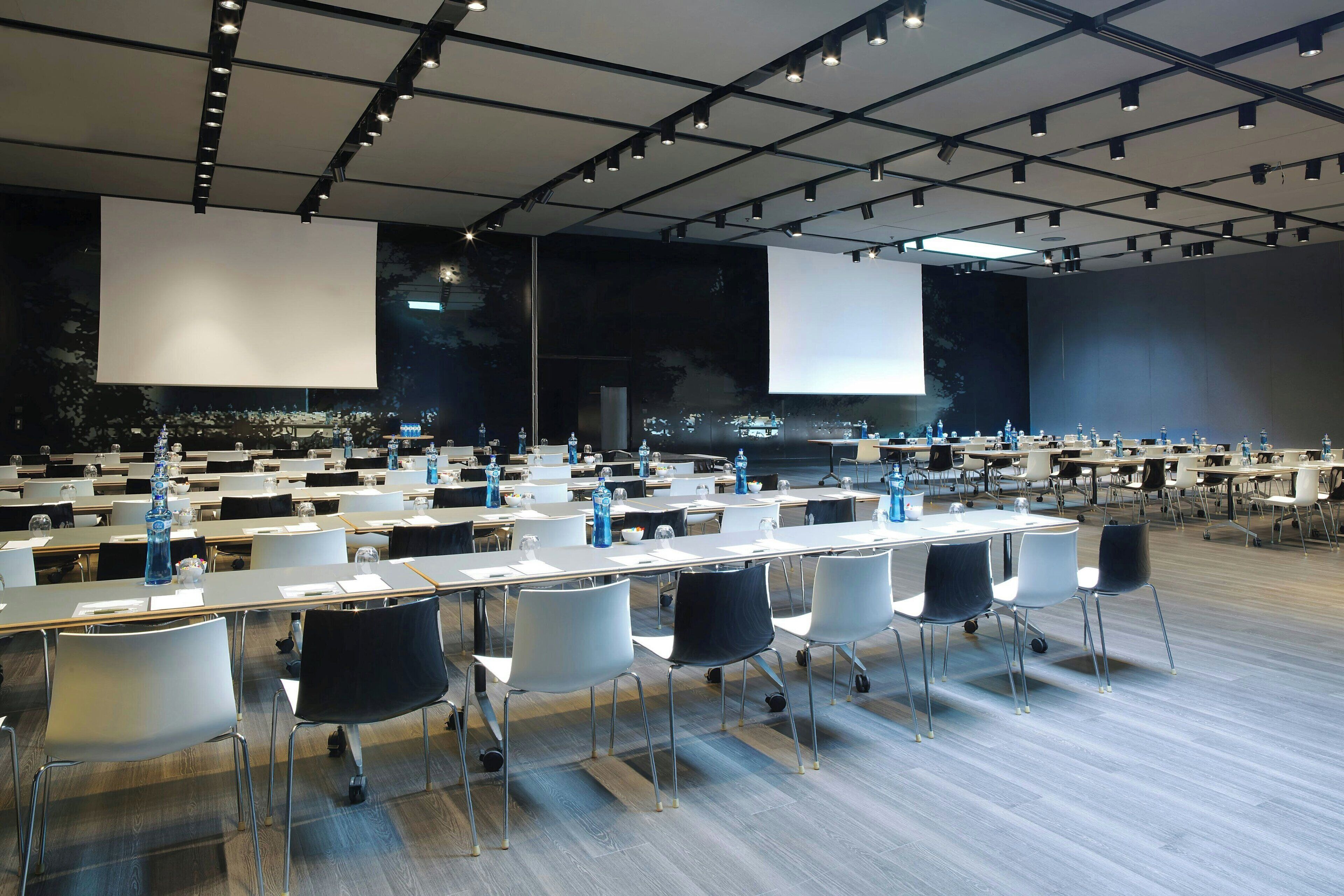 A conference room with rows of tables and chairs, large screens, and a cityscape view.
