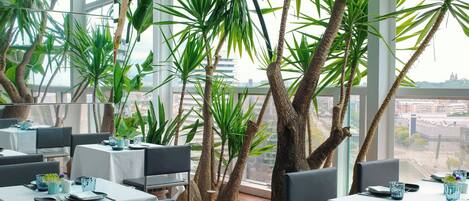 A restaurant with tables set for dining, surrounded by large indoor plants and a view of the cityscape through large windows.