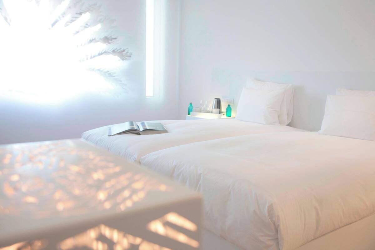 Ein ordentlich eingerichtetes Schlafzimmer mit einem großen Bett, einem Nachttisch mit einem Buch und einem Fenster mit Tageslicht.