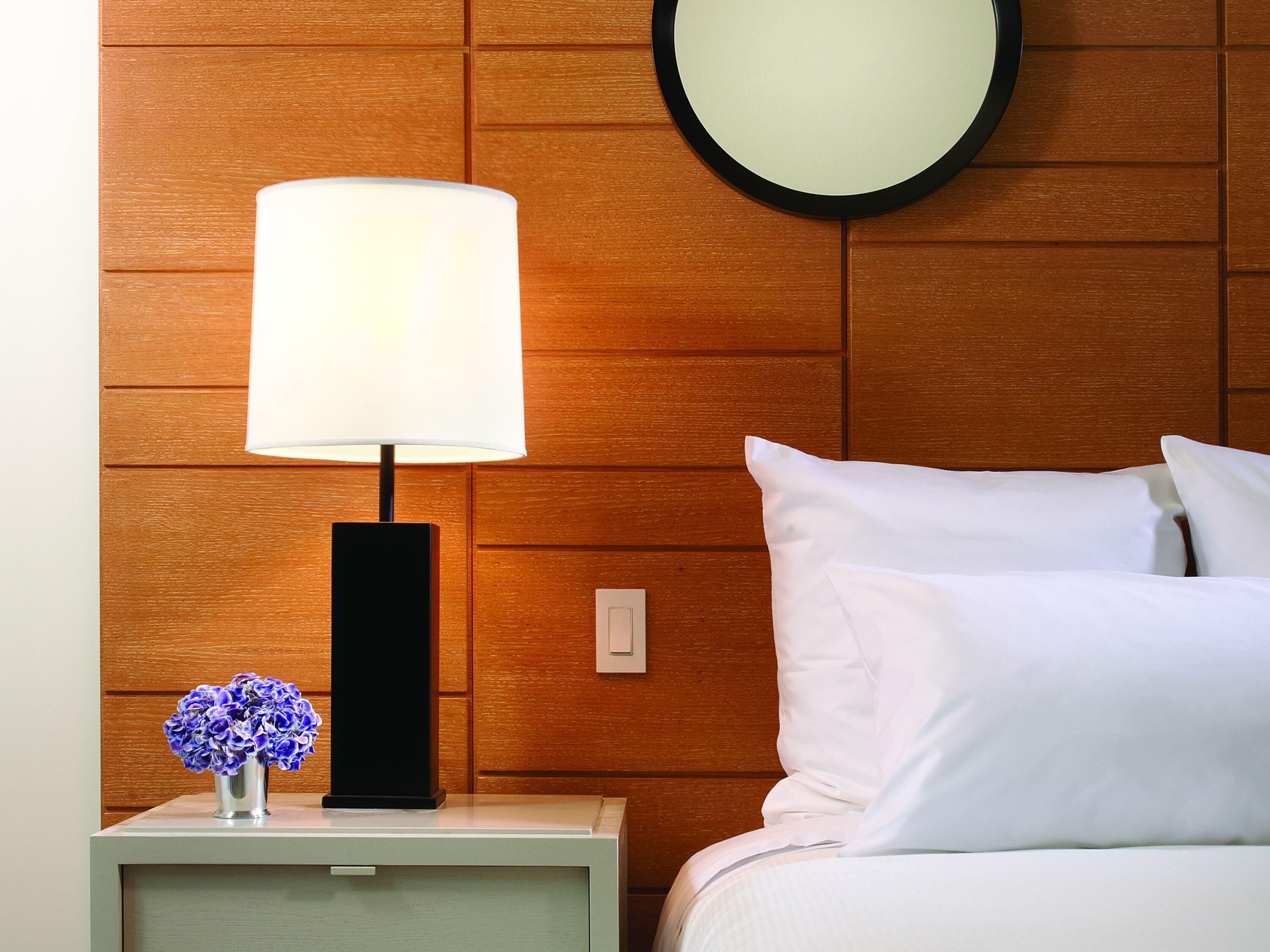 A neatly arranged hotel room with a bedside lamp, a wall-mounted round mirror, a nightstand with a vase of flowers, and a bed with white bedding.