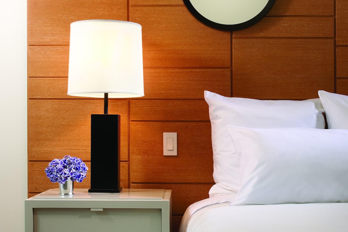 A neatly arranged hotel room with a bedside lamp, a wall-mounted round mirror, a nightstand with a vase of flowers, and a bed with white bedding.