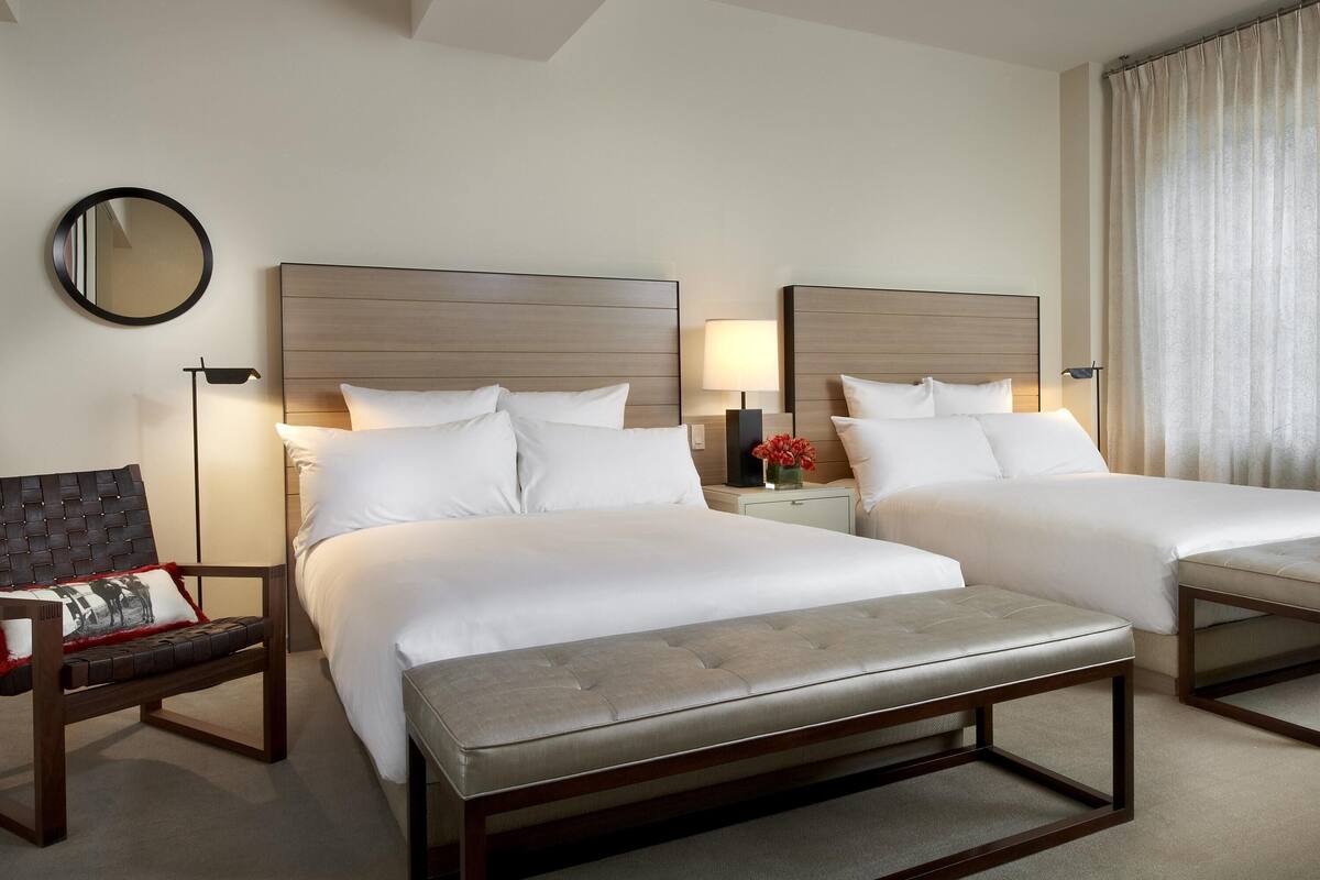 Habitación de hotel con dos camas, un banco, una silla, una mesita de noche con lámpara, un espejo y una ventana con cortinas.