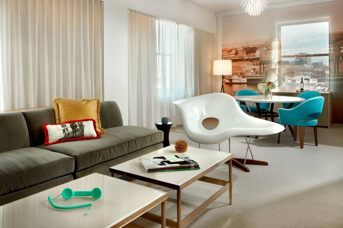 Una habitación de hotel moderna con sofá, una mesa de centro, zona de comedor con sillas y vistas a un paisaje urbano a través de amplios ventanales.