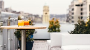 A table with a glass of orange juice on a balcony overlooking a cityscape with buildings and a tower.