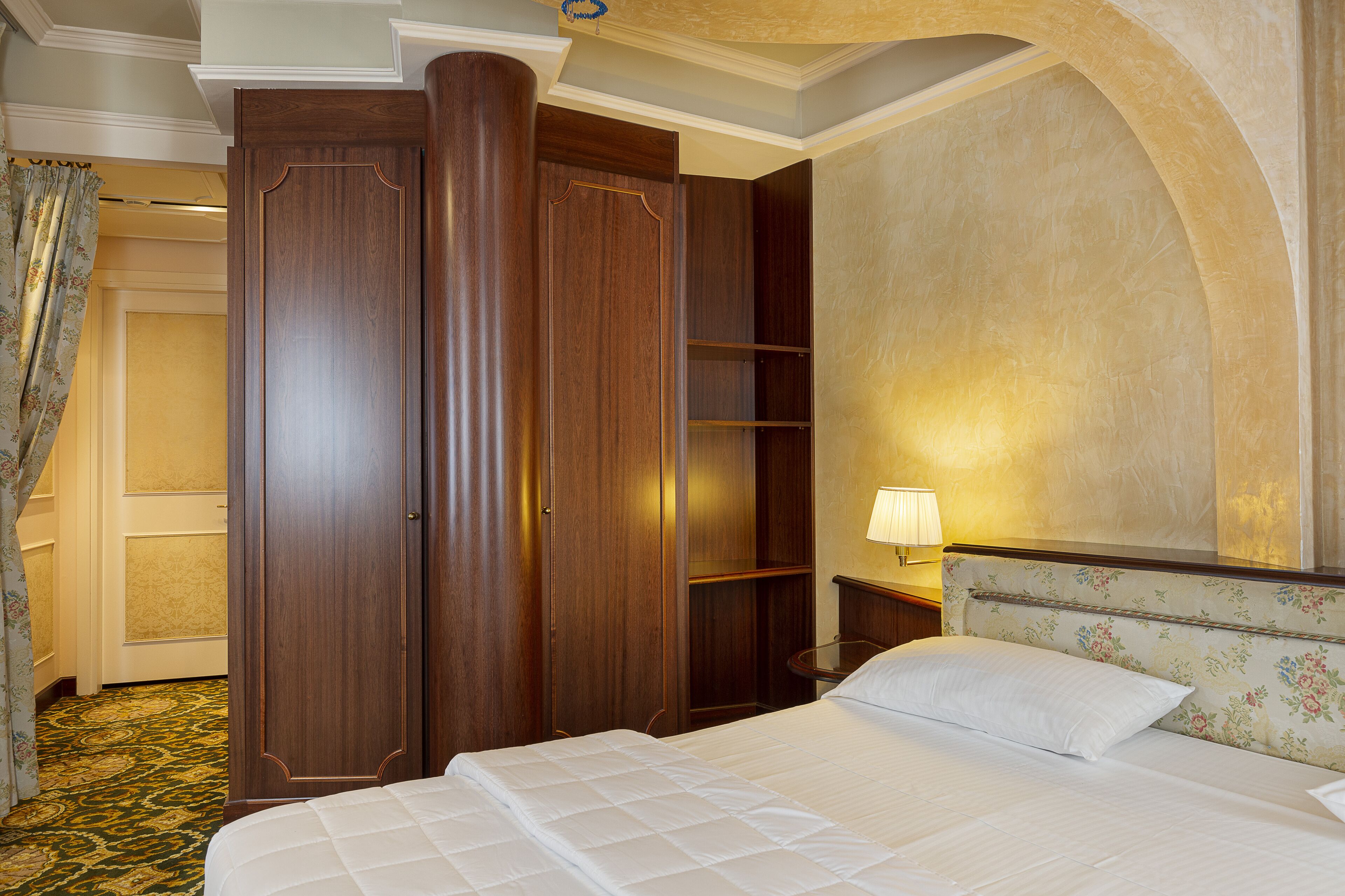 Ein Hotelzimmer mit einem großen Bett, einem Kleiderschrank, einem Nachttisch mit Lampe und einem blumengemusterten Kopfteil.