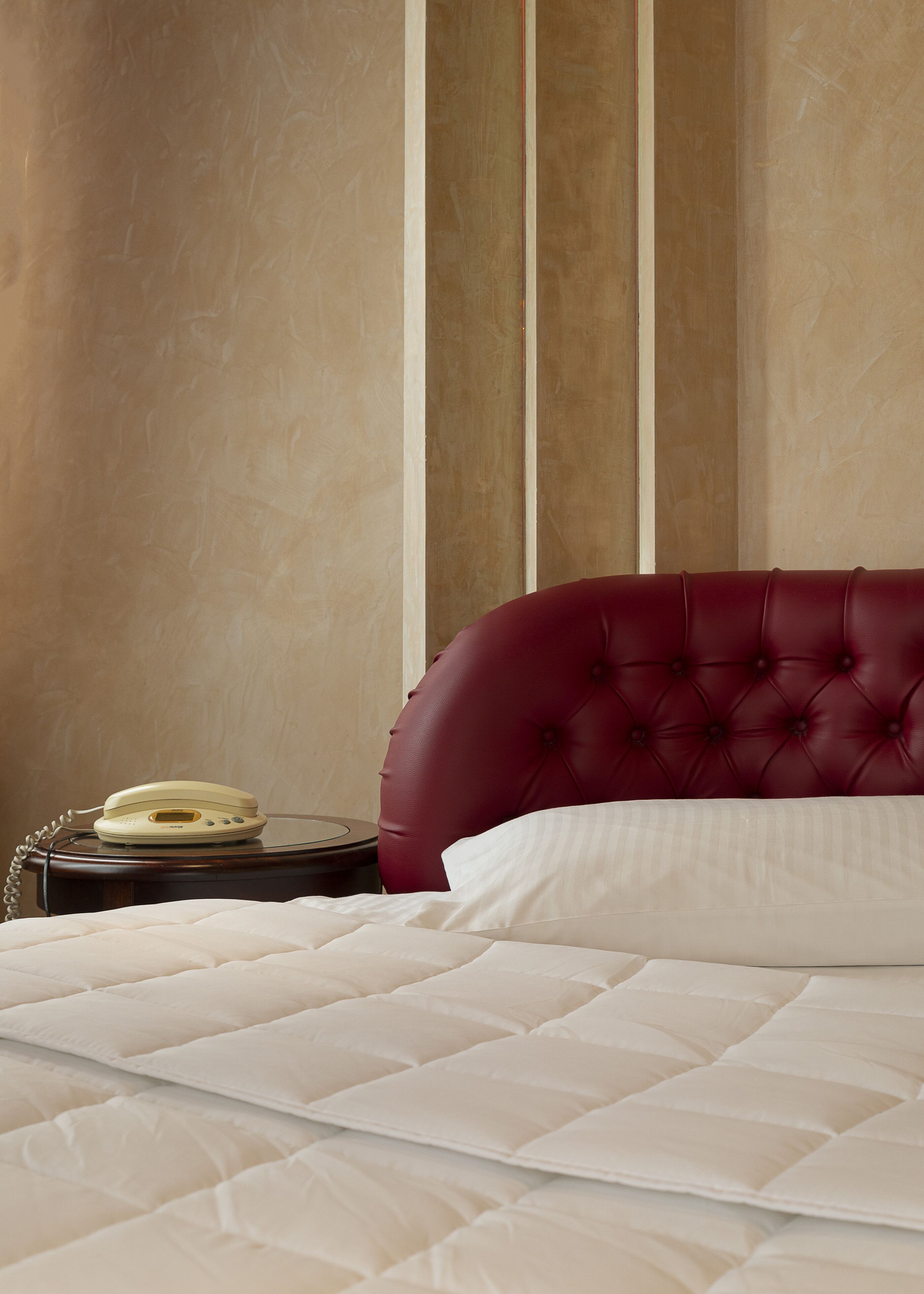 A bed with a red upholstered headboard, a white quilted mattress, and a bedside table with a cream-colored telephone.