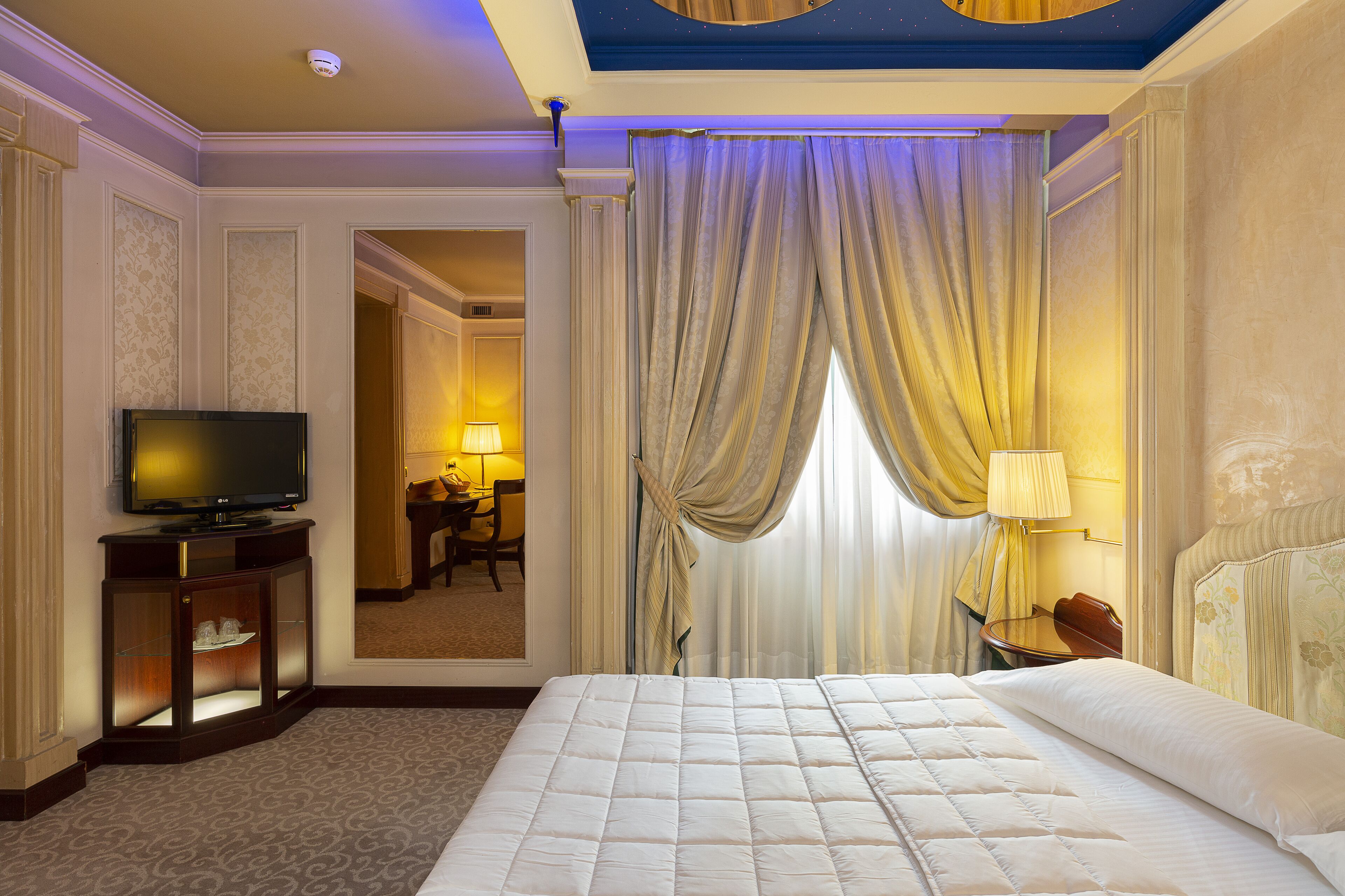 A hotel room with a large bed, a television on a stand, a desk with a lamp, and a view of another room through a door.