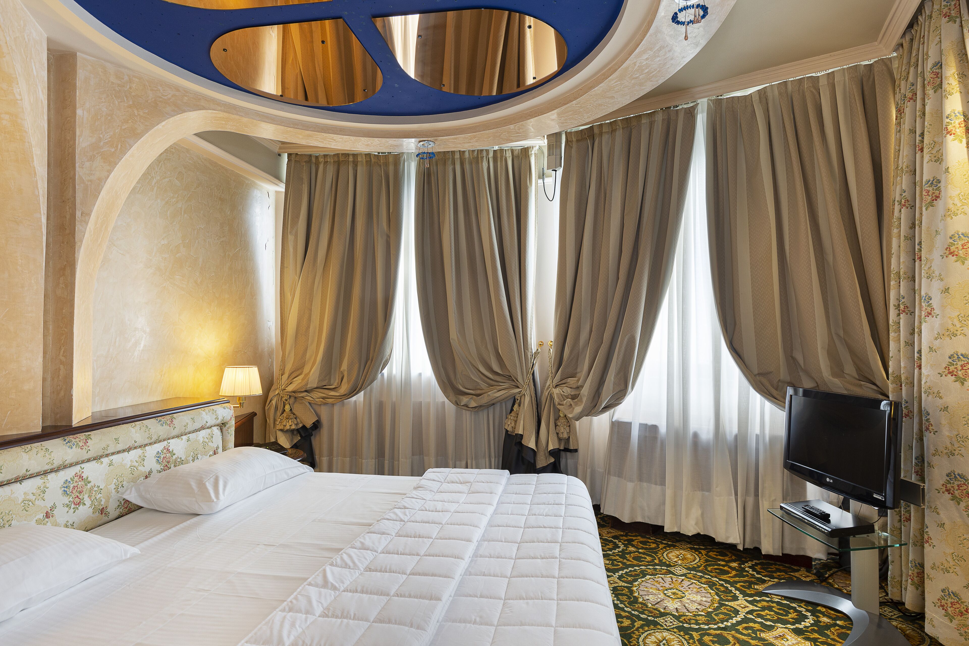 A hotel room with a bed, curtains, a television, and a patterned carpet.