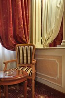 A wooden chair with a striped cushion, a round wooden table with a glass top, and a window with red and cream curtains.