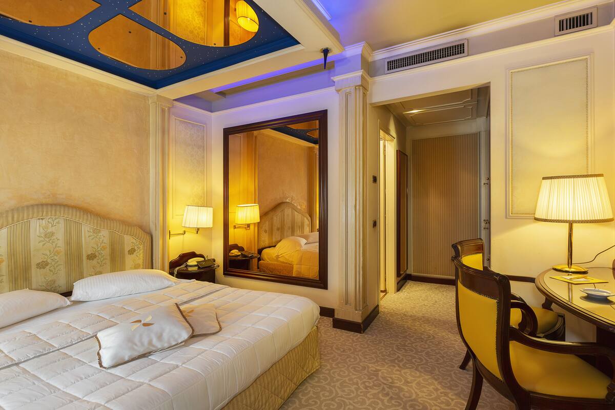 A hotel room with a large bed, a desk with a chair, and a mirror reflecting the room.
