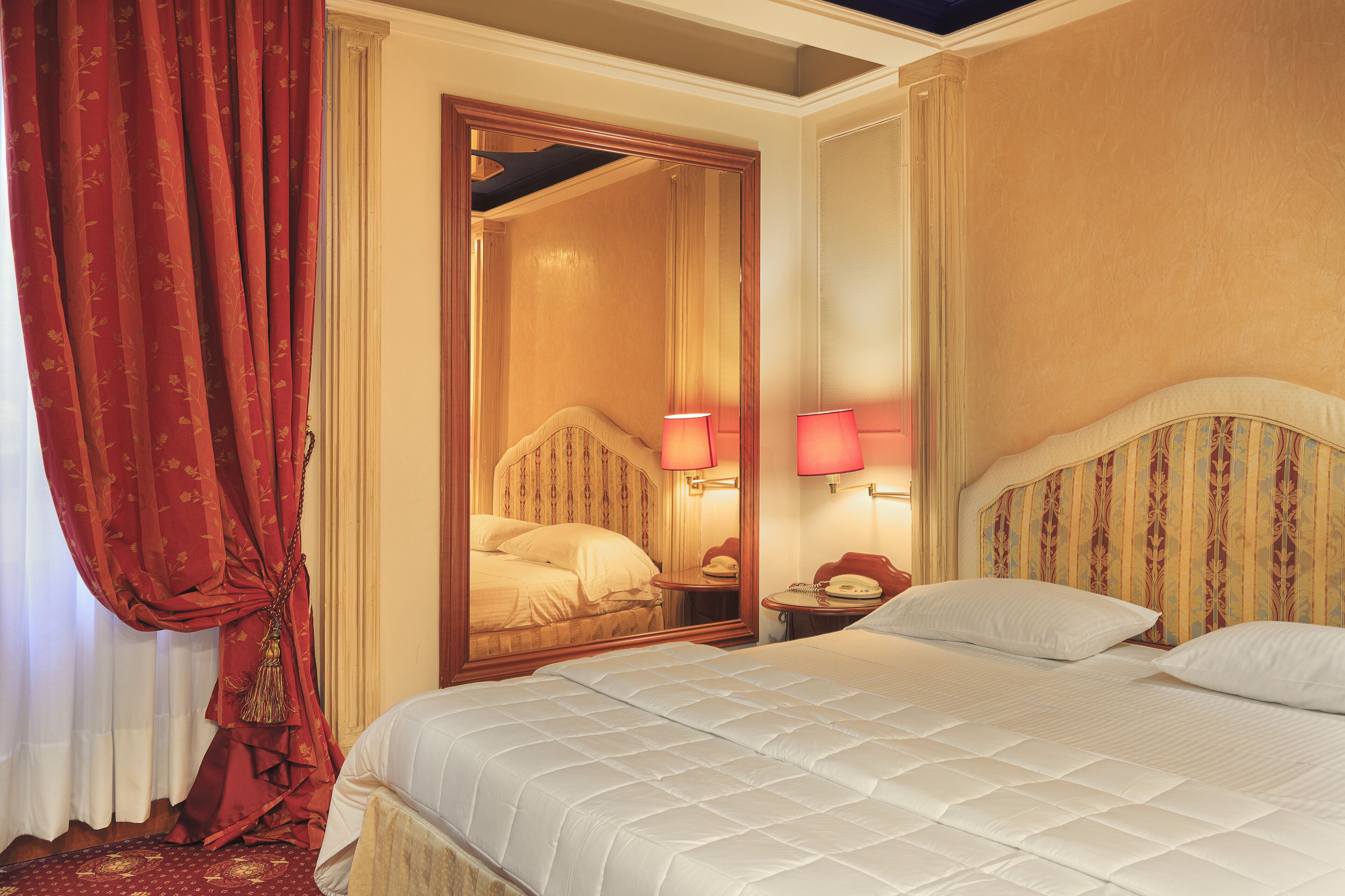 Una habitación de hotel con una cama grande, un espejo, dos lámparas de noche y una cortina roja.