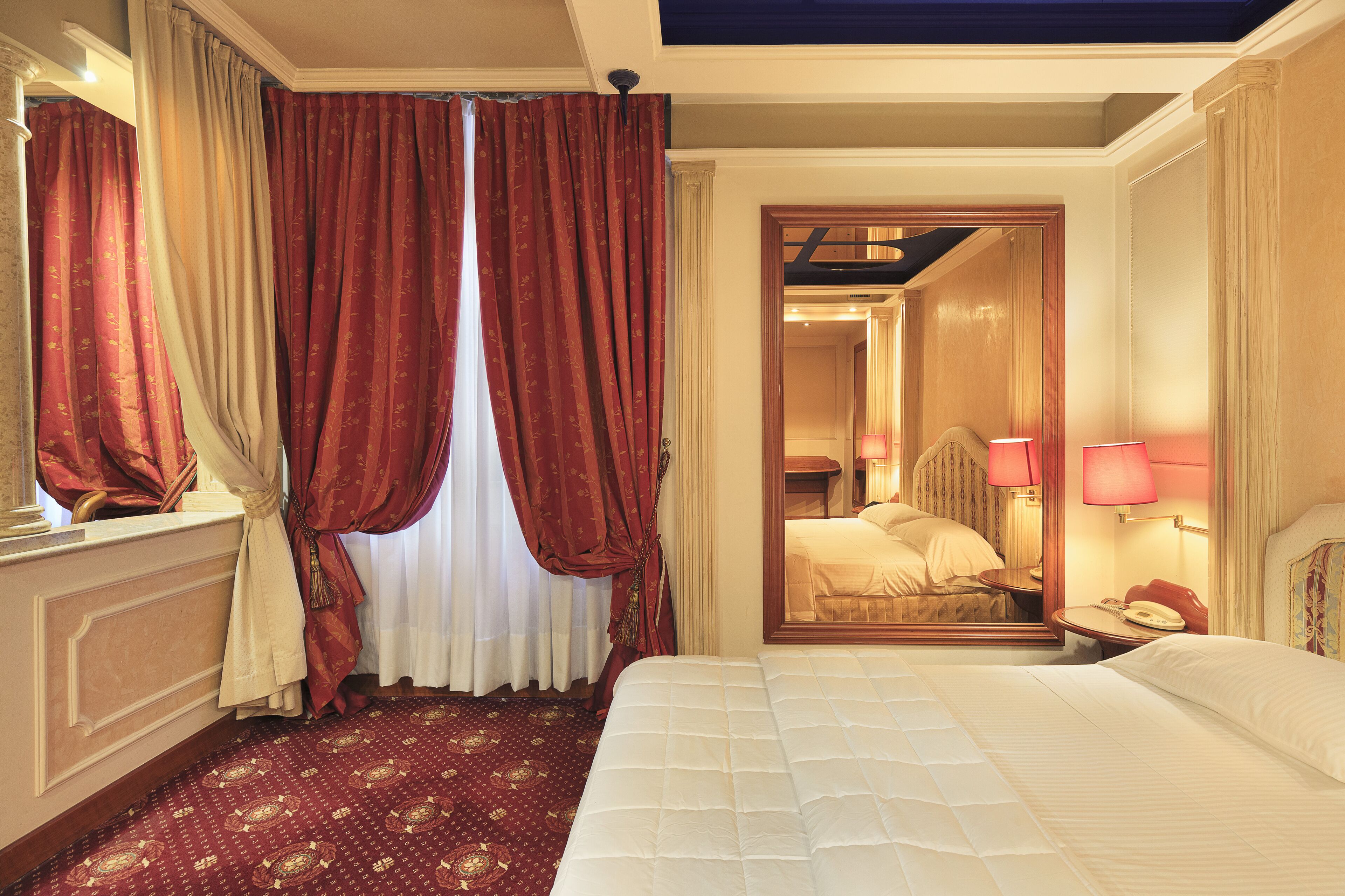 Camera d'albergo con un letto grande, uno specchio, un comodino con una lampada e un pavimento con tappeto rosso.