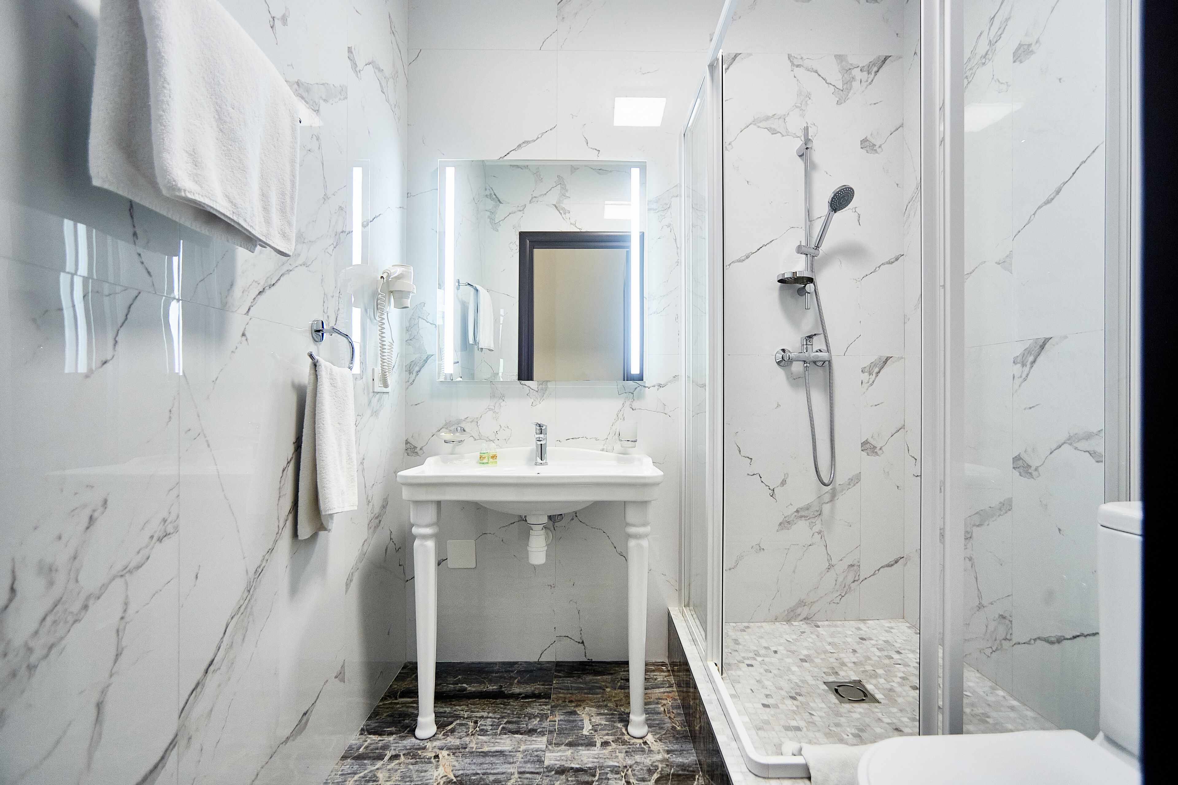 Une salle de bain avec un lavabo sur pied blanc, un miroir au-dessus, une douche avec une porte en verre et des murs à motifs de marbre.