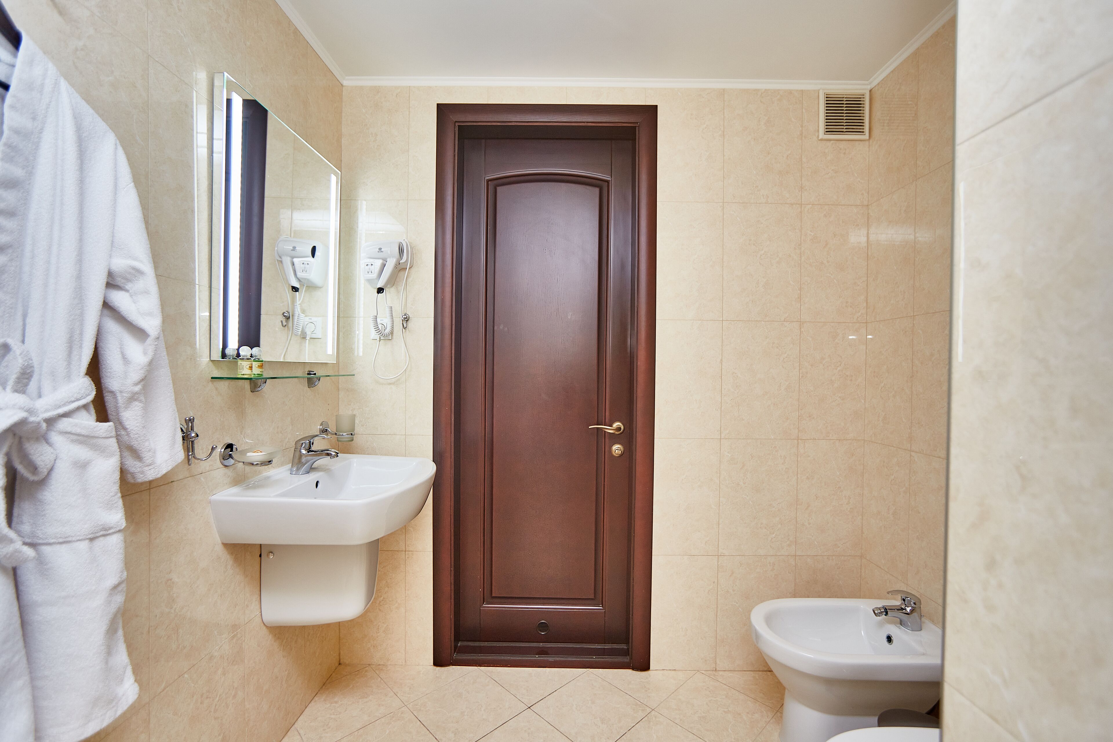 Un baño con una puerta marrón, un lavamanos blanco, un bidé y una bata colgada en la pared.