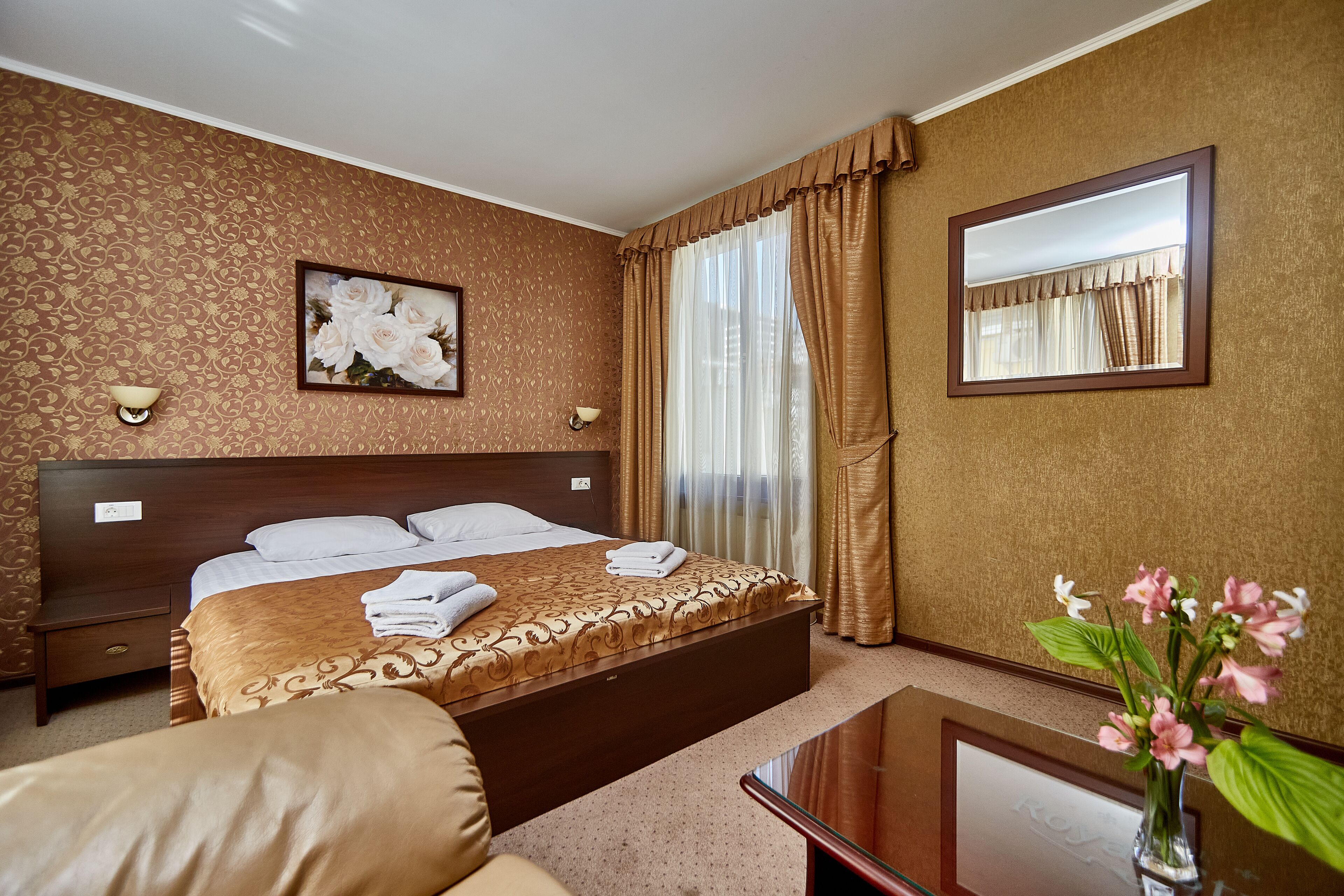 Una habitación de hotel con una cama grande, una mesita de noche, un espejo y un florero con flores.
