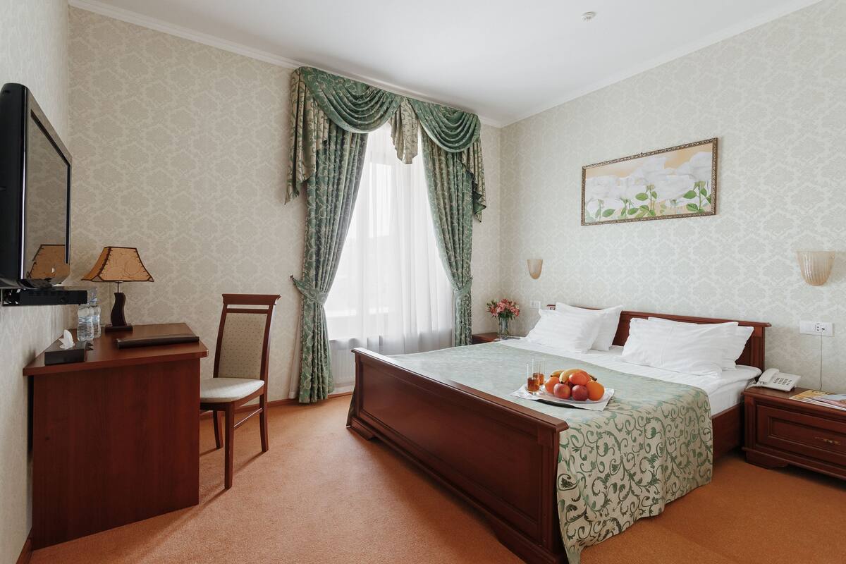 A hotel room with a wooden bed, a desk with a chair, a bedside table, a lamp, and a painting on the wall.