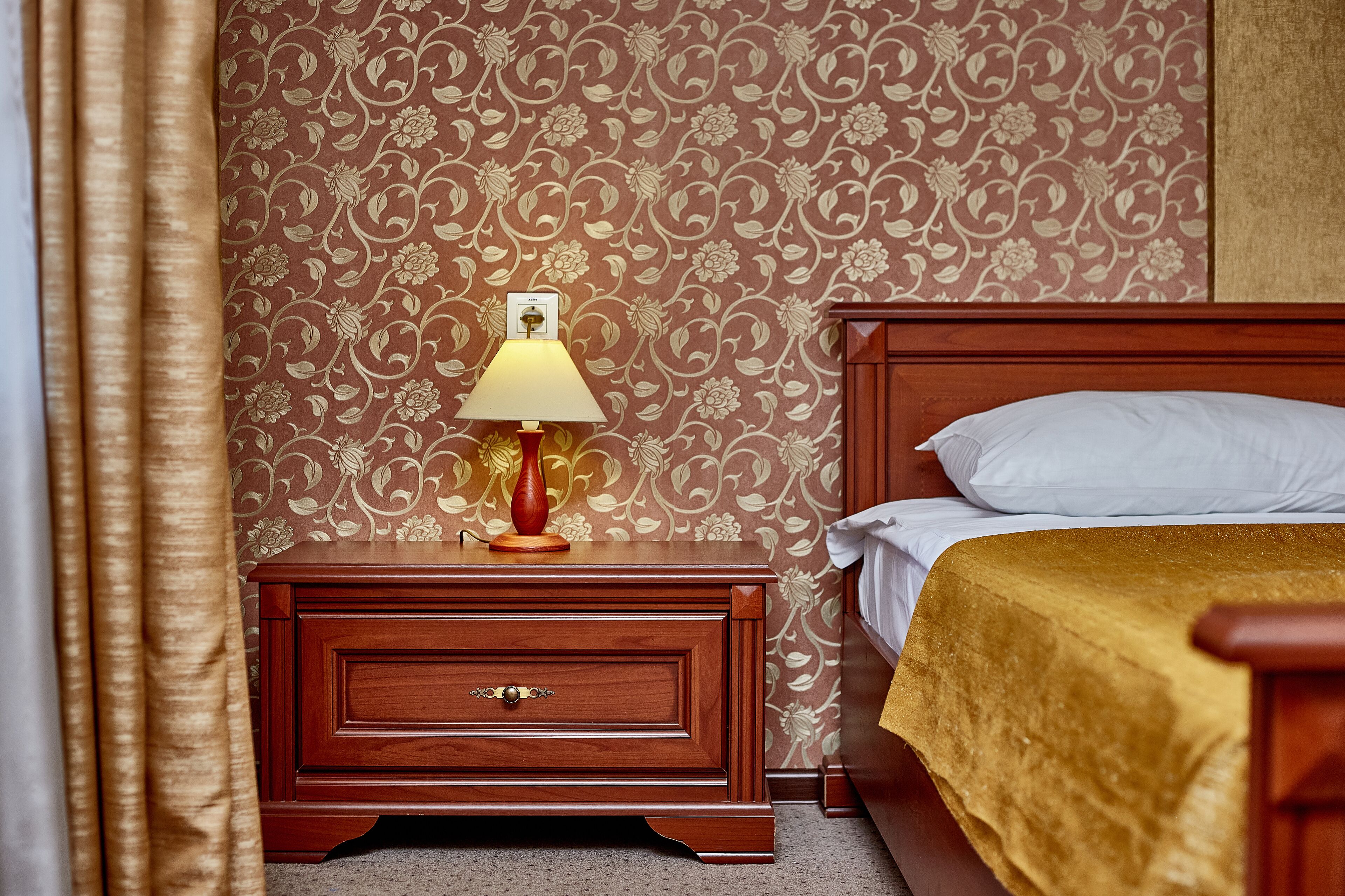 A hotel room with a wooden nightstand, a lamp, a bed with a gold bedspread, and a floral wallpaper pattern.