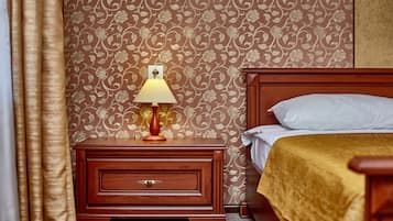 A hotel room with a wooden nightstand, a lamp, a bed with a gold bedspread, and a floral wallpaper pattern.