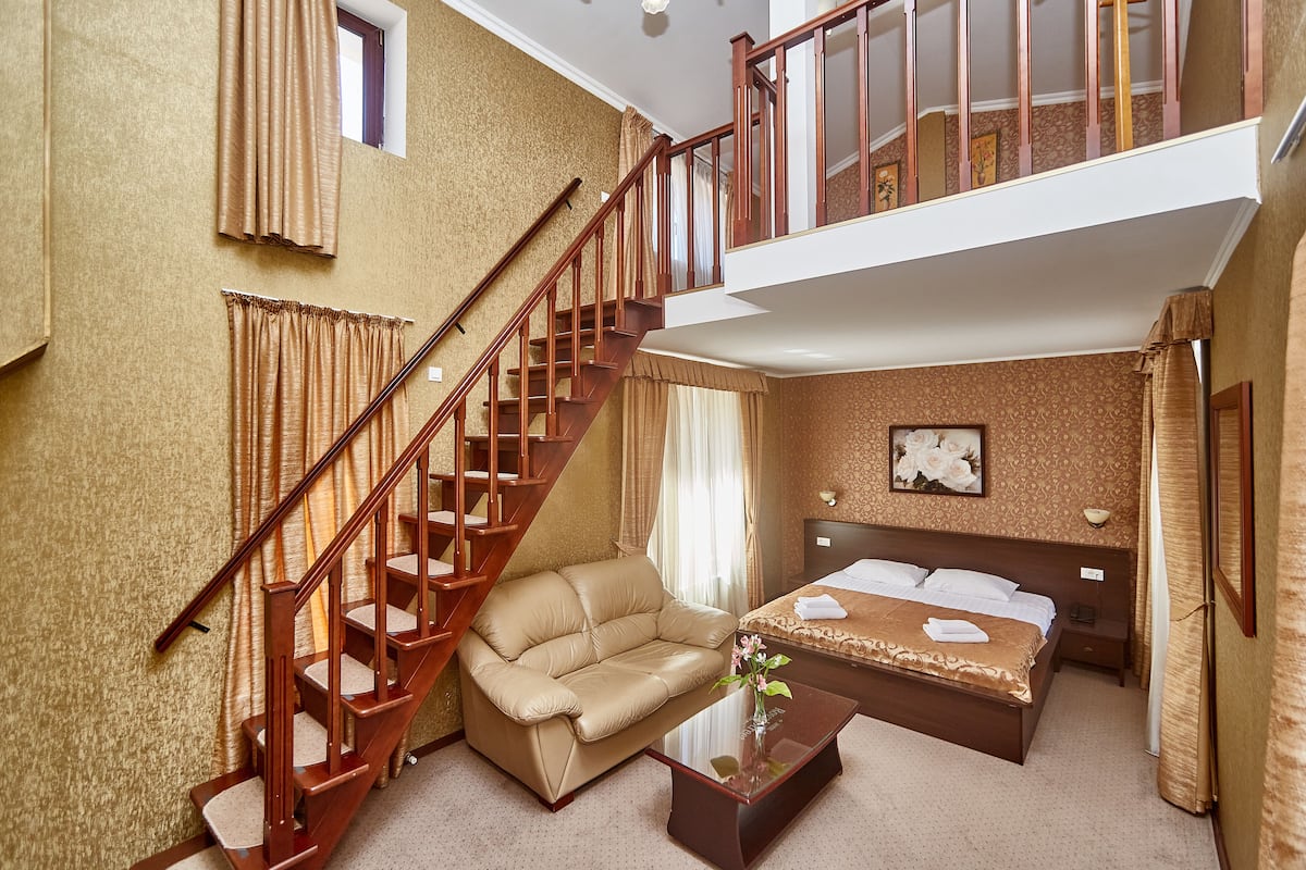 A hotel room with a sofa, a bed, a wooden staircase, and a view of the upper floor.