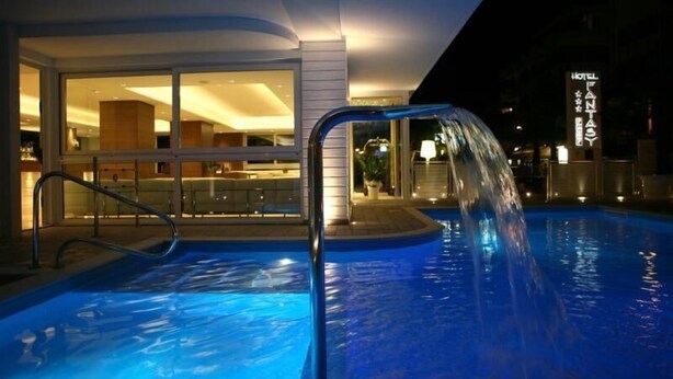 A swimming pool with a water feature, a glass door entrance to a building, and a sign for 'Hotel Fantasy'. - Hotel Fantasy (Riccione)