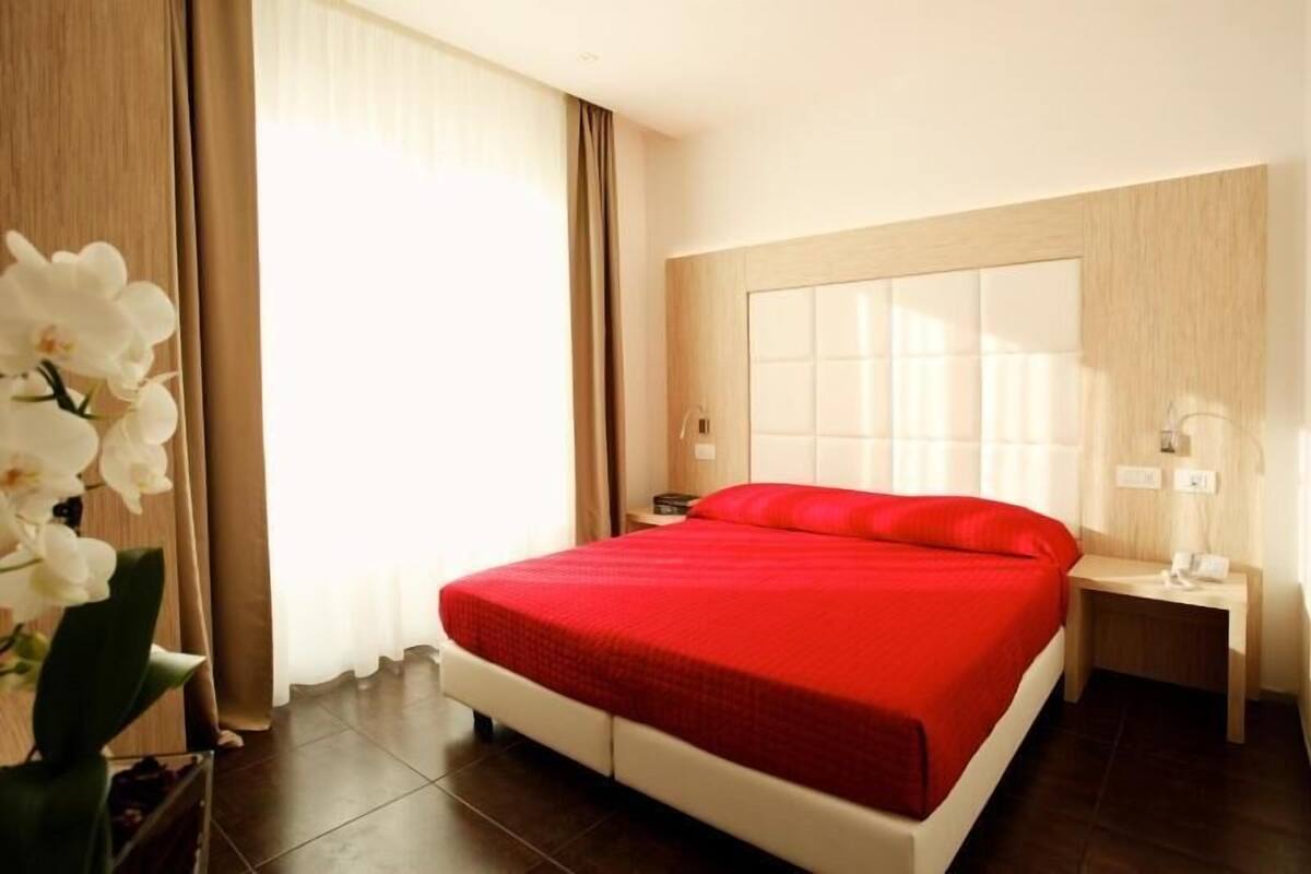 A hotel room with a large bed, a bedside table, a nightstand, a window with curtains, and a potted orchid.