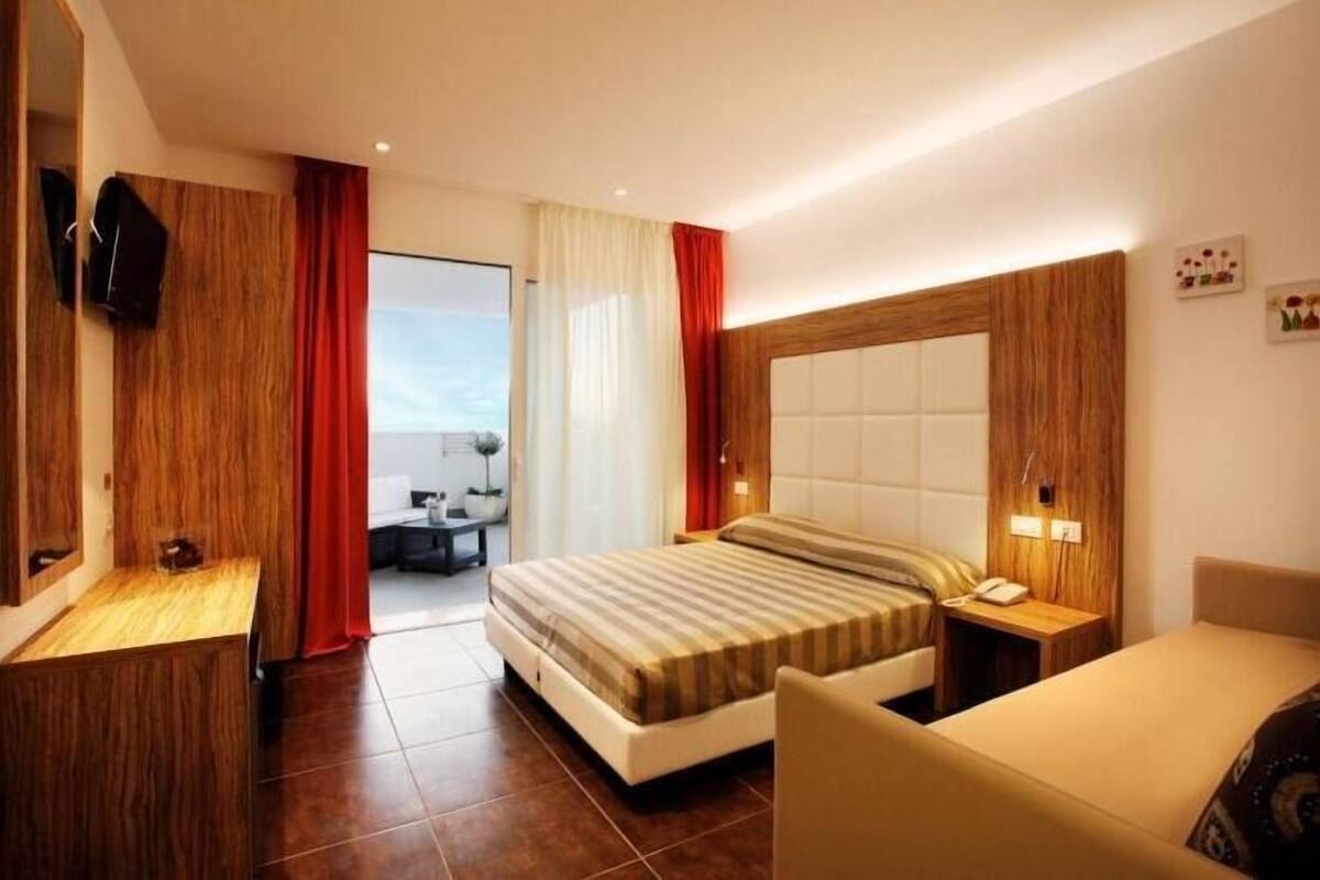 A modern hotel room with a large bed, a bedside table, a TV mounted on the wall, and a view of the outdoors through the window.