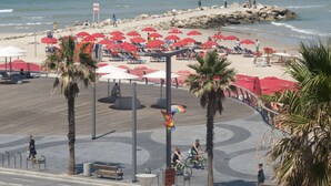 A beachside promenade with red umbrellas, palm trees, and a rocky shoreline. - TLV 88 Sea Hotel (Tel Aviv)