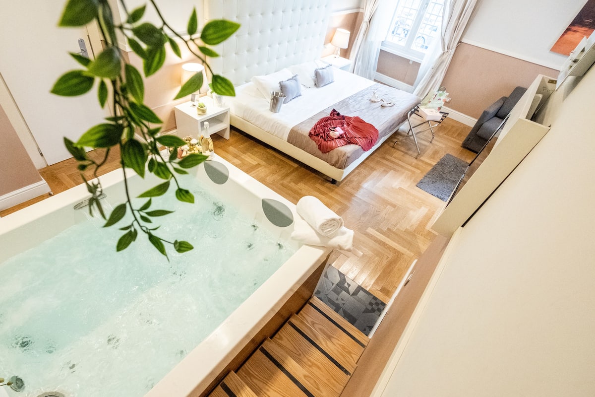 A modern bedroom with a large bed, a bedside table, a nightstand, a window with curtains, and a bathtub with a towel.