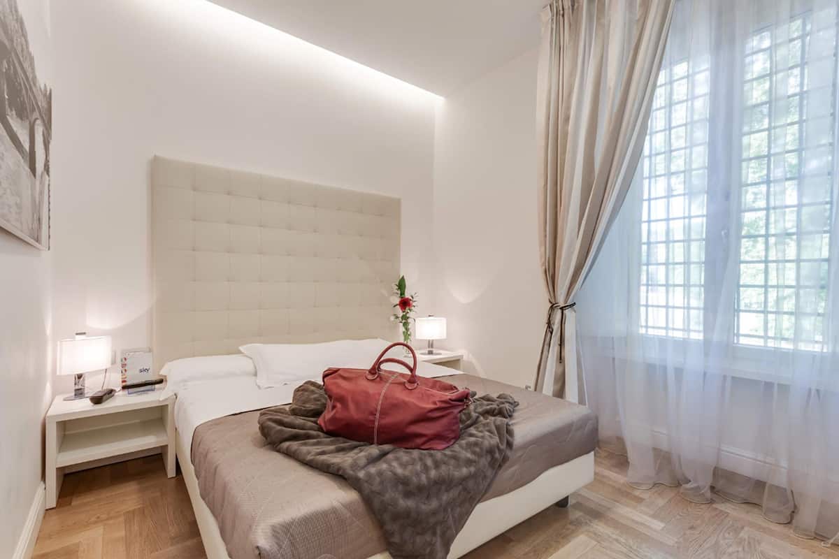 A hotel room with a large bed, two bedside tables with lamps, a red handbag on the bed, and sheer curtains on the window.