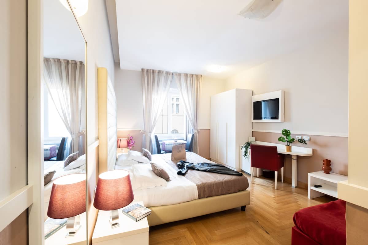 A modern hotel room with a bed, bedside table, lamp, TV, desk, chair, and a view of the city.