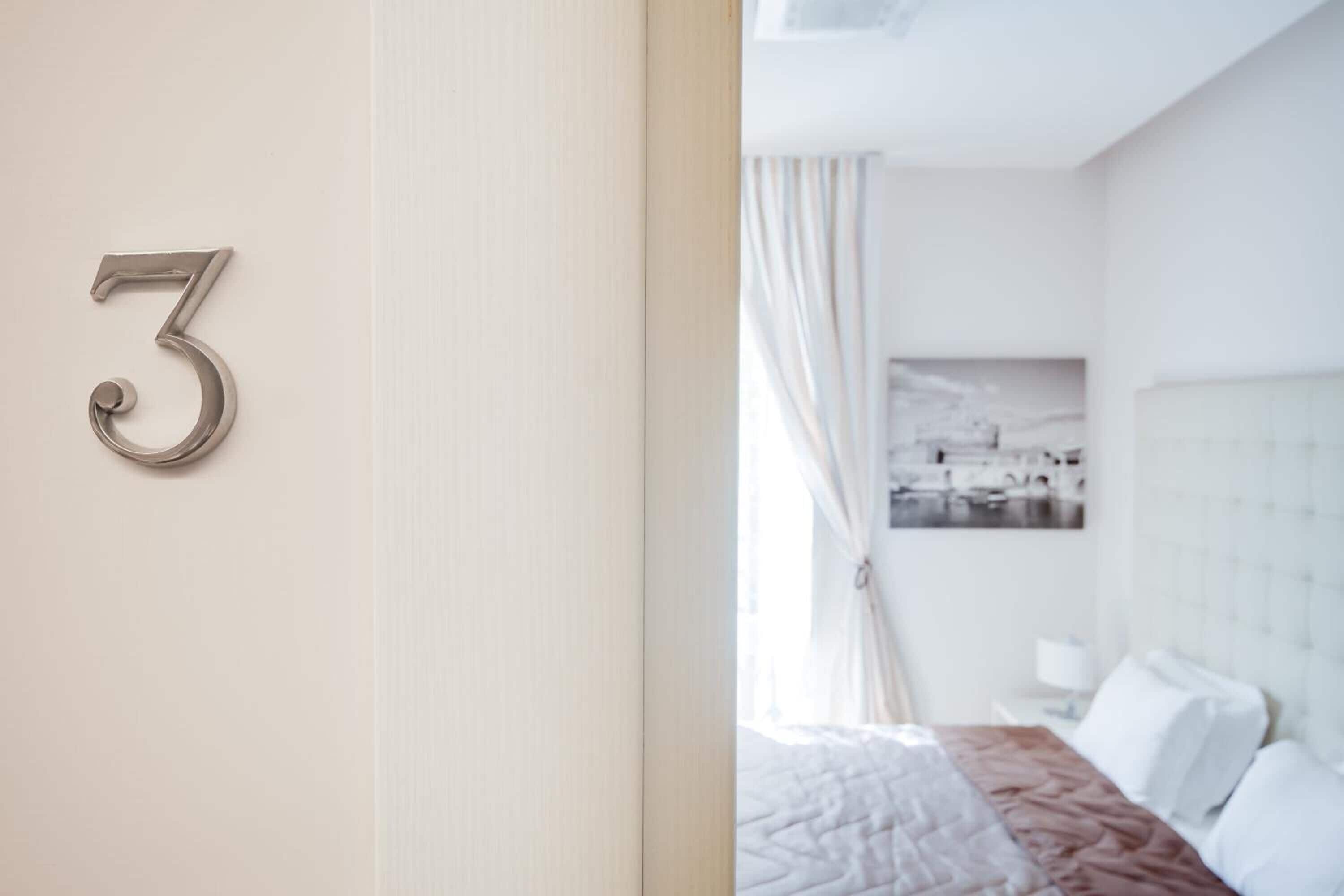 A hotel room with a large number '3' on the wall, a bed with white bedding, a headboard, and a window with curtains.