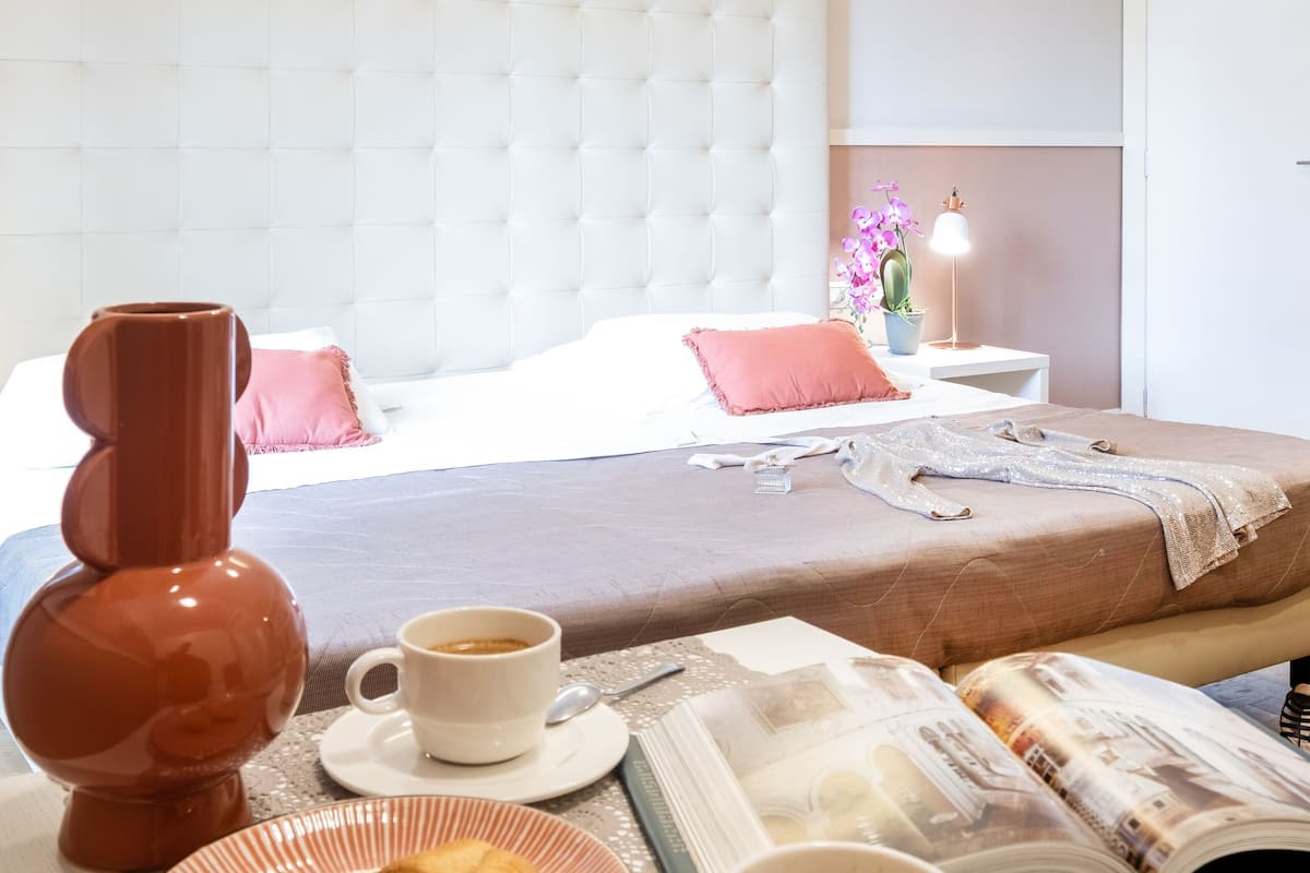 A neatly arranged hotel room with a bed, a bedside table with a lamp, a coffee setup, and a magazine.