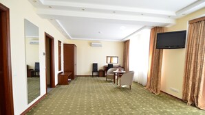 Ein Hotelzimmer mit grünem Teppich, einem flachbildfernseher an der Wand, einem Schreibtisch mit Stuhl und einem großen Spiegel.