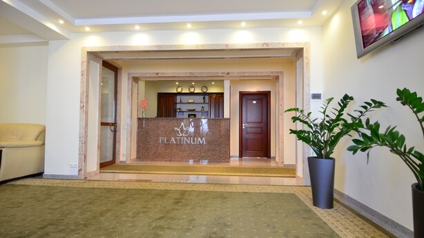 Ein Hotel-Empfangsbereich mit einem 'Platinum'-Schild, einem Empfangstresen, einem Fernsehbildschirm und einer Zimmerpflanze.
