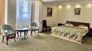 Ein Hotelzimmer mit einem Bett, zwei Sessel, einem kleinen Tisch und einem Stuhl. An den Wänden hängen zwei Bilder und ein Fenster mit Vorhängen.