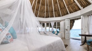 Een slaapkamer met een bed bekleed met een witte hemel, uitzicht op de oceaan en een rieten dak.