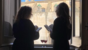 Two people reading by a window with a view of a building.