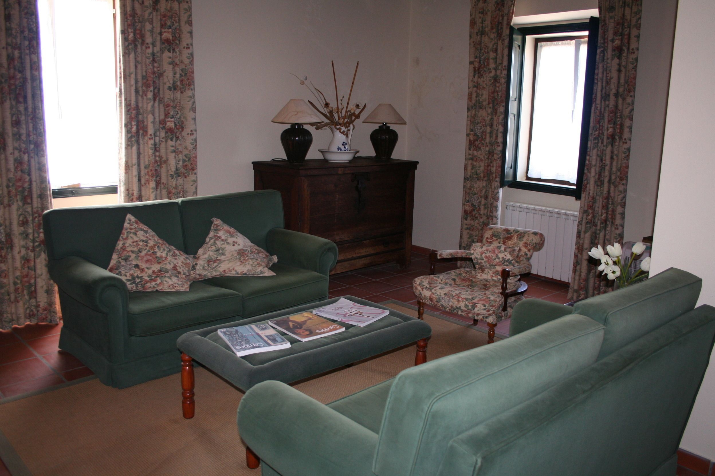 Un salon avec un canapé vert, une table basse sur laquelle sont posés des magazines, un fauteuil à motifs floraux et un meuble en bois avec des lampes.