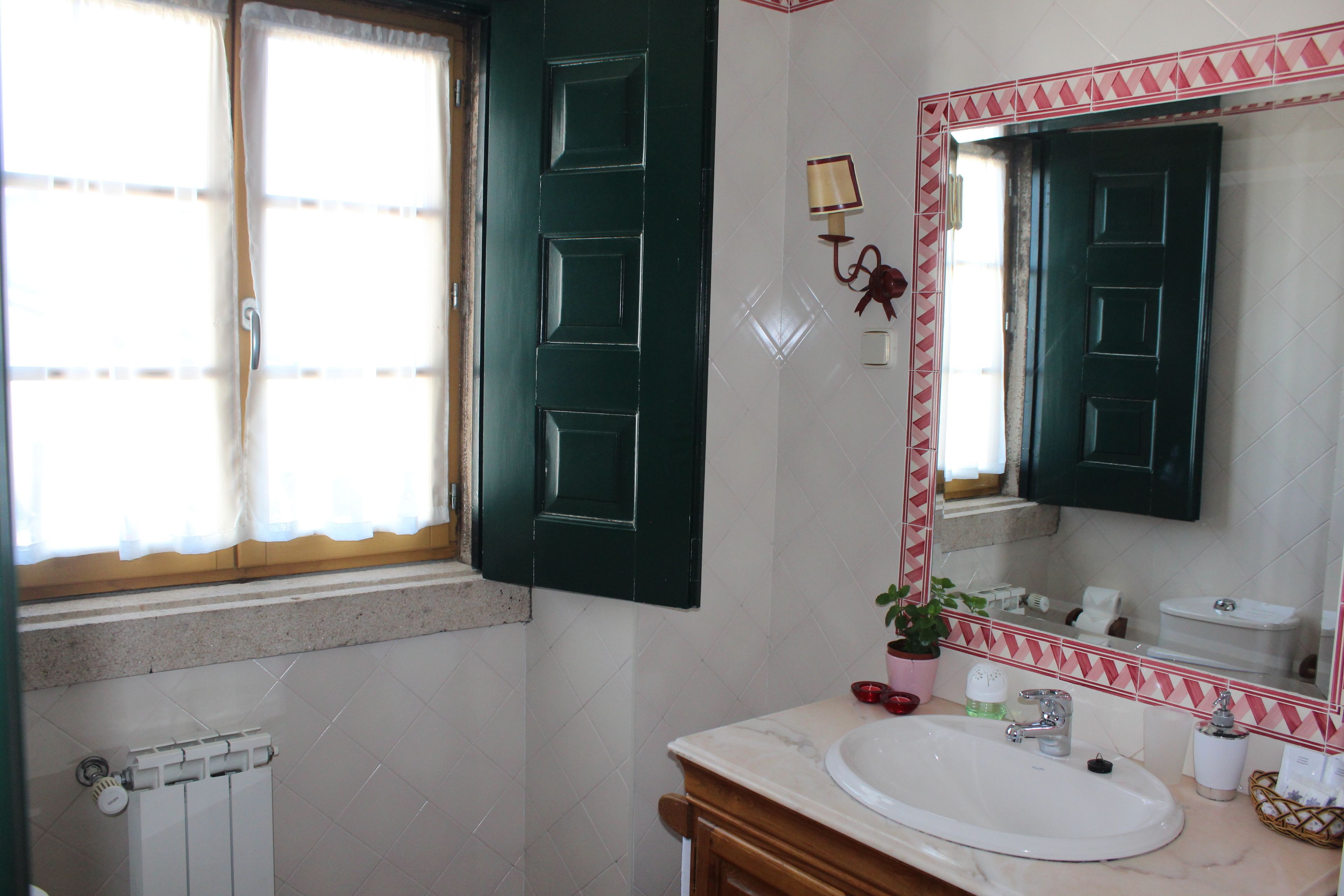 A bathroom with a white sink, a mirror with a red and white frame, a potted plant, and a window with curtains.