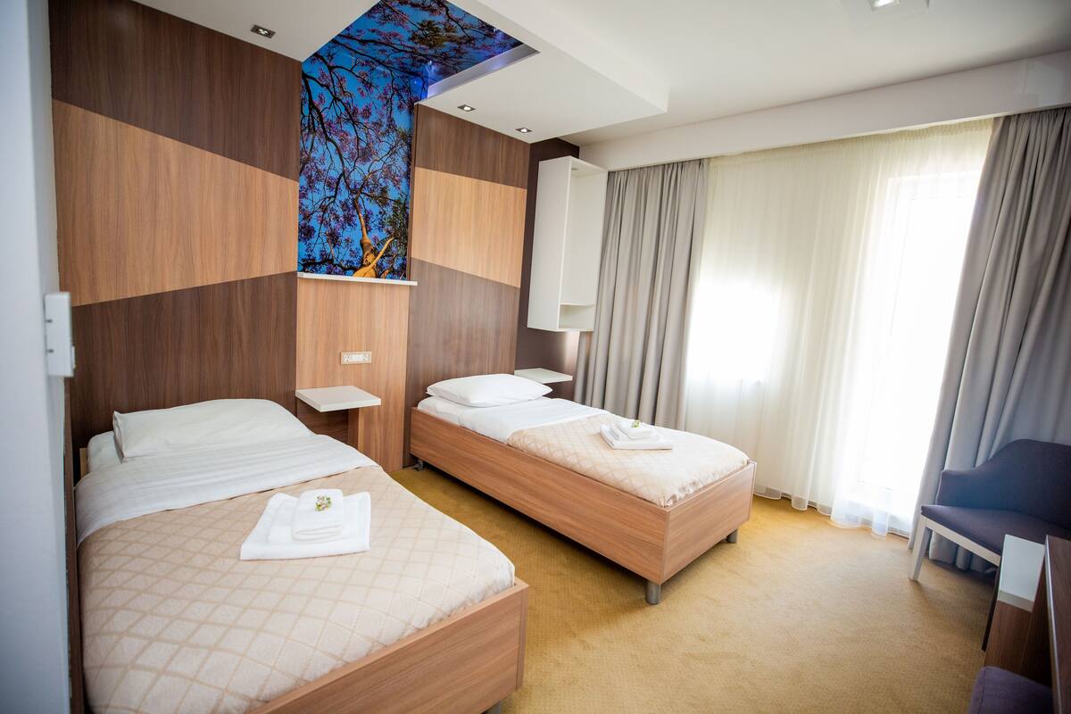 A hotel room with two beds, a large window with curtains, a wooden wall panel, and a ceiling with a tree mural.