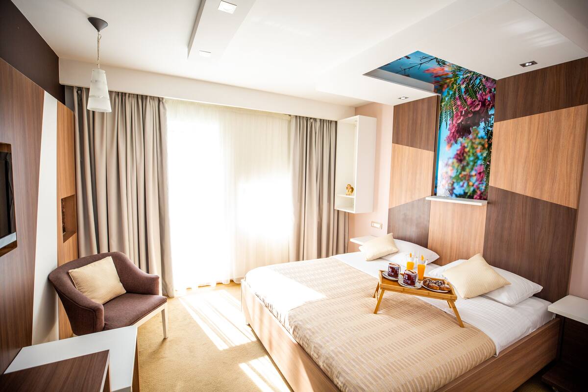 A modern hotel room with a large bed, a sitting area with a chair, a small table with breakfast items, and a wall-mounted TV.