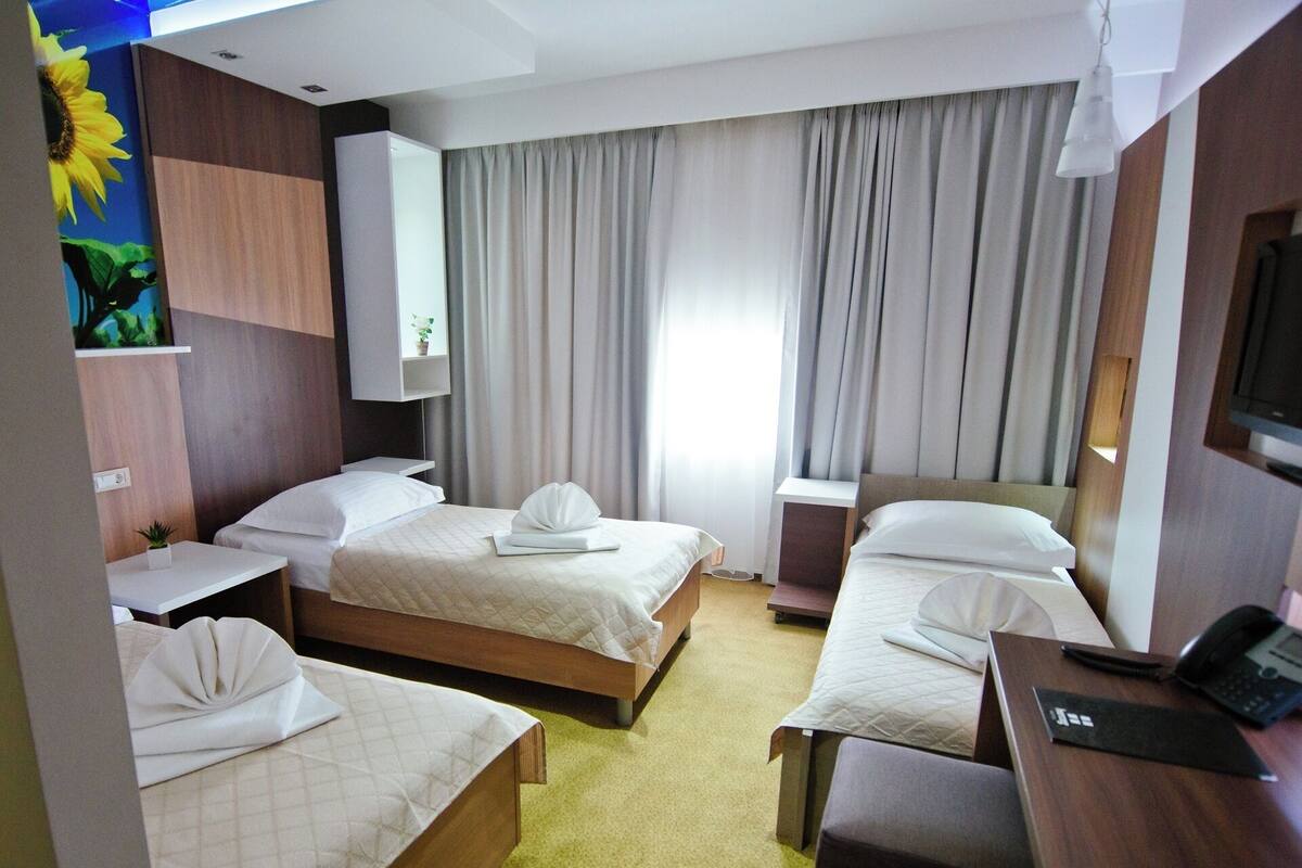 A hotel room with two beds, a desk, a chair, a TV, and a large window with curtains.