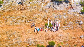 Paisaje rocoso con árboles dispersos y un grupo de personas reunidas alrededor de una cruz blanca.