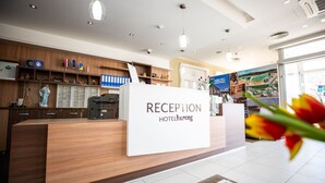 Un mostrador de recepción con un letrero que dice "RECEPTION HOTEL HERCEG". Hay estantes con varios objetos y un ventanal grande al fondo.