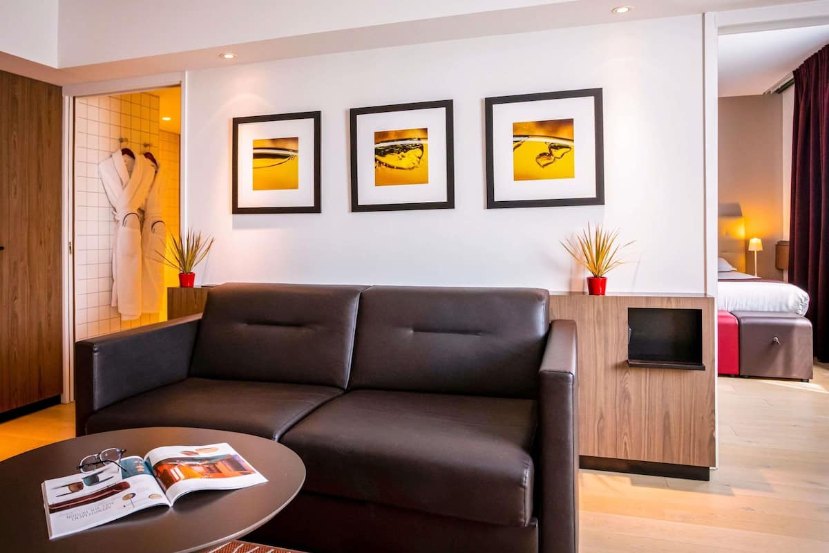 A modern hotel room with a leather sofa, a small coffee table, and a bed with a red suitcase. There are three framed pictures on the wall and a wooden cabinet.