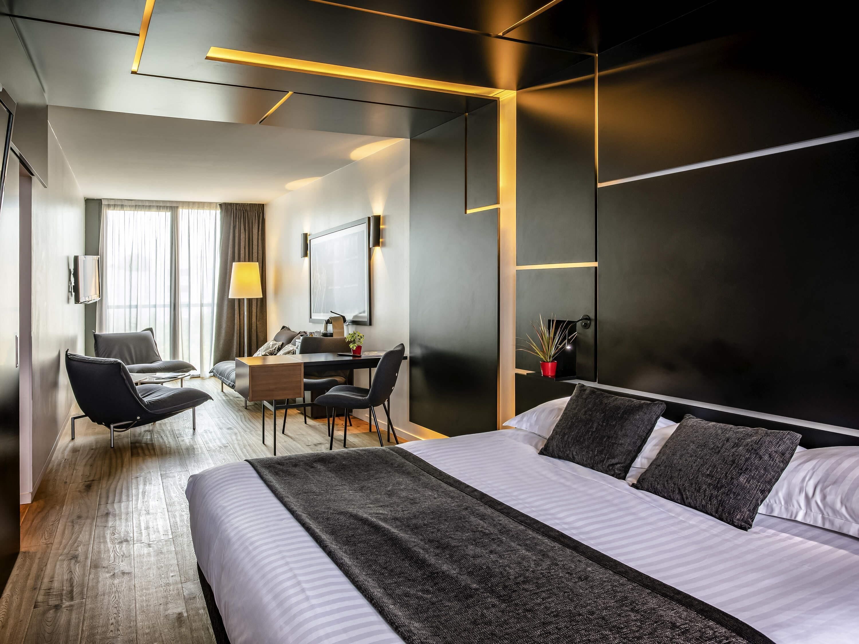 Une chambre d’hôtel moderne avec un grand lit, un bureau, deux chaises, une télévision et une fenêtre avec des rideaux.