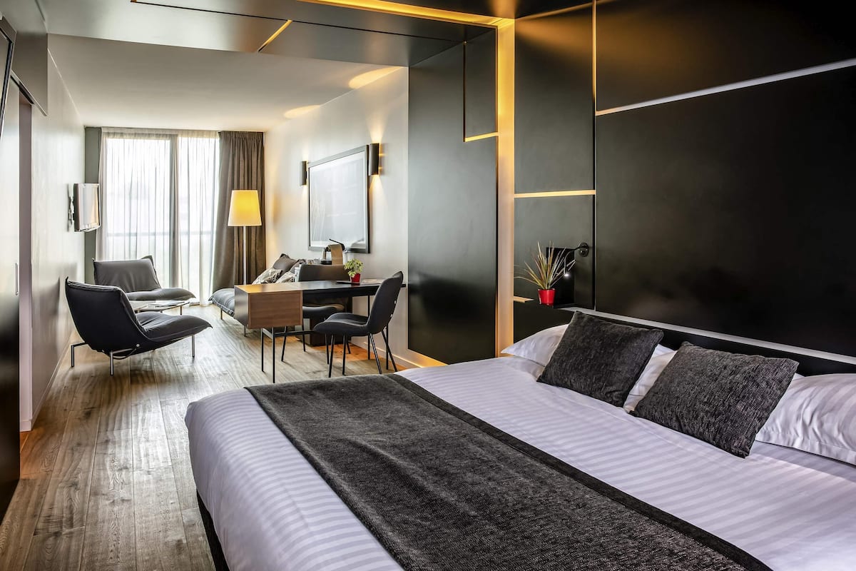 Une chambre d’hôtel moderne avec un grand lit, un bureau, deux chaises, une télévision et une fenêtre avec des rideaux.