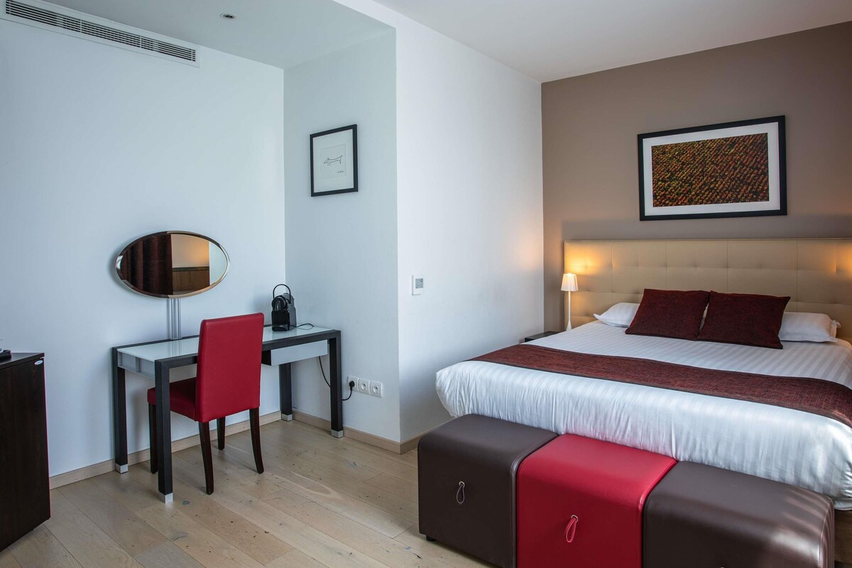 A hotel room with a bed, a desk, a chair, a mirror, and a wall-mounted picture.