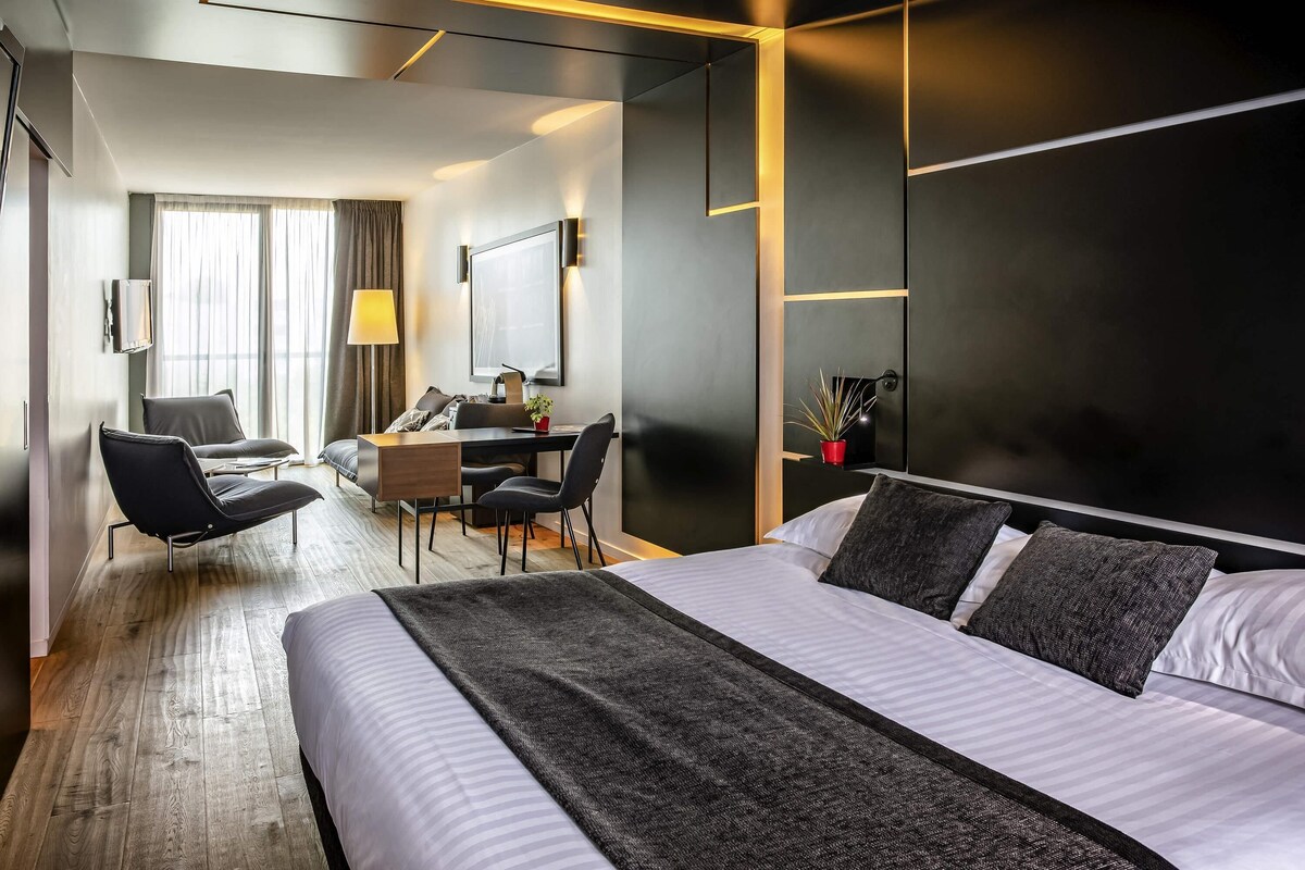 A modern hotel room with a large bed, a desk, two chairs, a TV, and a window with curtains.